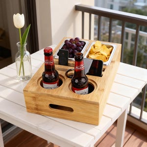 May include: A bamboo serving tray with two beer bottles, two food compartments, and two phone slots. The tray holds grapes and potato chips. A small handle is cut into the side. The tray sits on a white table.