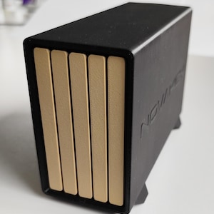 Könnte beinhalten: Ein schwarzes rechteckiges Gerät mit fünf vertikalen beigefarbenen Paneelen auf der Vorderseite. Auf der Seite des Geräts ist das Wort "NOVIA" eingeprägt. Das Gerät hat ein modernes, minimalistisches Design.