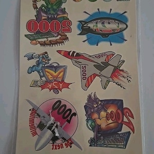Puede incluir: Una lámina de tatuajes temporales con varios gráficos de dibujos animados. Los diseños incluyen un avión, un dirigible, un ordenador y criaturas alienígenas. Los tatuajes están empaquetados en una funda de plástico transparente con las palabras "TEMPORARY TATTOOS".