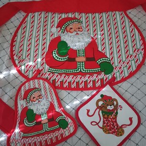 Puede incluir: Un juego de cocina con temática navideña que incluye un delantal, un guante de horno y un salvamanteles. El delantal y el guante tienen un diseño de Papá Noel sobre un fondo a rayas rojas y blancas. El salvamanteles presenta un oso en un diseño de calcetín.