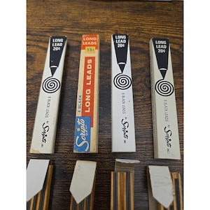 May include: Four vertical rectangular boxes of Scripto long leads, each containing six black leads. Three boxes are white with black text and graphics, while one is orange, white, and blue. Each box displays the text Long Lead and Scripto.