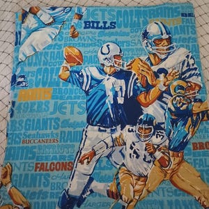 以下が含まれることがあります： アメフト選手が活躍する様子を描いた水色の生地。Colts、Rams、Giantsなどのチーム名がデザインされ、様々なユニフォームを着た選手がボールを保持したり投げたりしています。背景にはチーム名が散りばめられています。