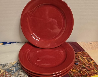 May include: A stack of four round, solid red ceramic plates. Each plate features a smooth, glossy finish with a slightly raised rim and a flat center. The plates are uniform in color and shape, stacked vertically to display their circular profile.