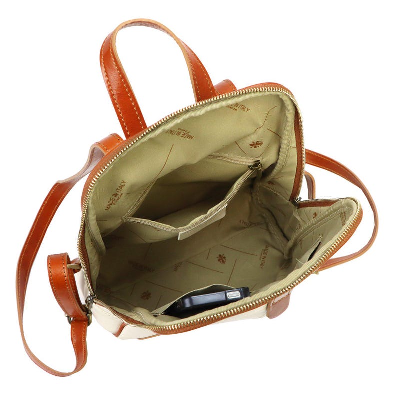 May include: Open view of a cream and brown leather backpack. The bag has a top handle and adjustable straps. The interior is beige with multiple pockets and a zipper closure. A black smartphone is visible inside the bag.