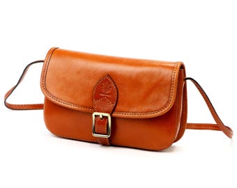 May include: A tan leather crossbody bag with a flap closure and a gold-coloured buckle. The bag has a long, thin strap and a stitched design around the edges. The bag is made of smooth leather.
