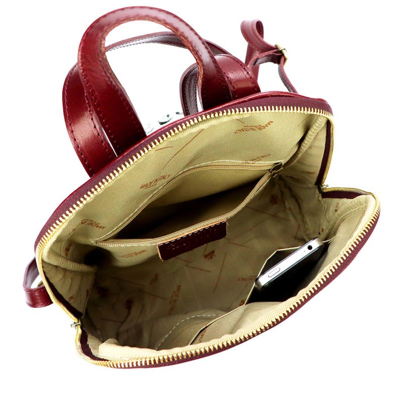 May include: Burgundy leather backpack with a top handle and adjustable straps. The interior features multiple pockets and a zippered compartment. A smartphone is visible inside the main compartment. The bag has a gold-colored zipper and a light tan interior lining.