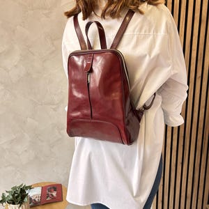 May include: A burgundy leather backpack with a square shape and a top handle. The backpack has a zipper closure and adjustable straps. The bag is worn over a white shirt and blue jeans.