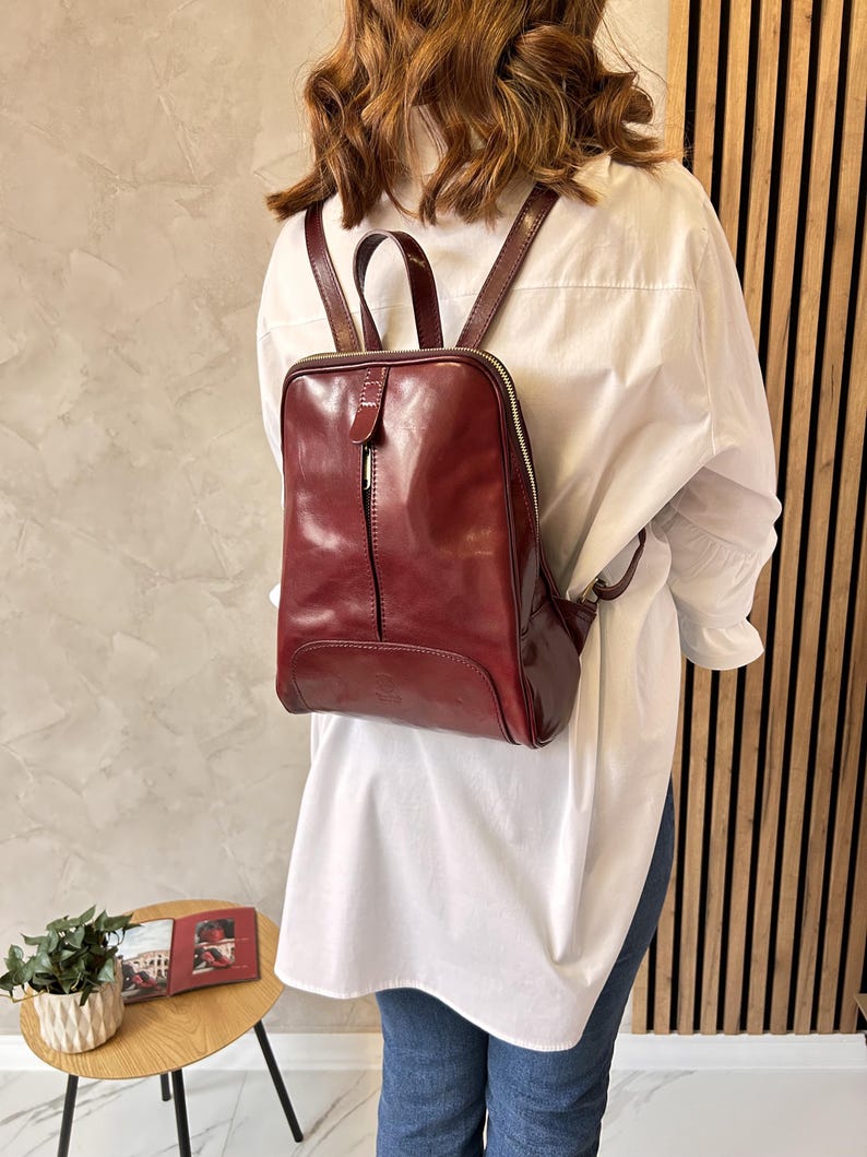May include: A burgundy leather backpack with a square shape and a top handle. The backpack has a zipper closure and adjustable straps. The bag is worn over a white shirt and blue jeans.
