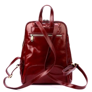 May include: A burgundy leather backpack with a glossy finish. The bag has a top handle, adjustable shoulder straps, and multiple zippered compartments. The hardware is gold-toned. The backpack is designed for everyday use.