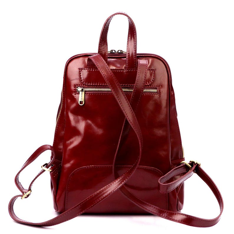 May include: A burgundy leather backpack with a glossy finish. The bag has a top handle, adjustable shoulder straps, and multiple zippered compartments. The hardware is gold-toned. The backpack is designed for everyday use.