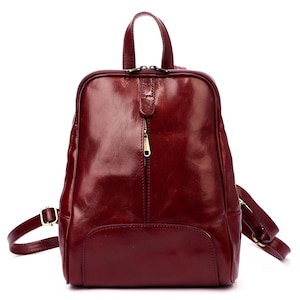 May include: A burgundy leather backpack with a top handle and adjustable shoulder straps. The backpack has a front pocket, a zipper closure, and gold-colored hardware. The bag is approximately 12 inches tall and 10 inches wide.