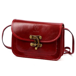 Könnte beinhalten: Eine rote Leder-Umhängetasche mit Klappenverschluss und Messingfarbenem Verschluss. Die Tasche hat einen langen, dünnen Riemen und ein kleines geprägtes Design auf der Vorderseite. Die Tasche ist etwa 20 cm breit und 15 cm hoch.