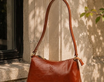 May include: A cognac-colored leather shoulder bag with a smooth finish, a single thin strap, and a top zipper closure. The front features a small embossed logo and text. The bag has a rectangular, structured shape with rounded bottom corners.