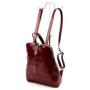 May include: A burgundy leather backpack with adjustable straps and a top handle. The bag features a zippered main compartment and a smaller front pocket. The hardware is gold-toned, and the bag is displayed on a silver metal stand.