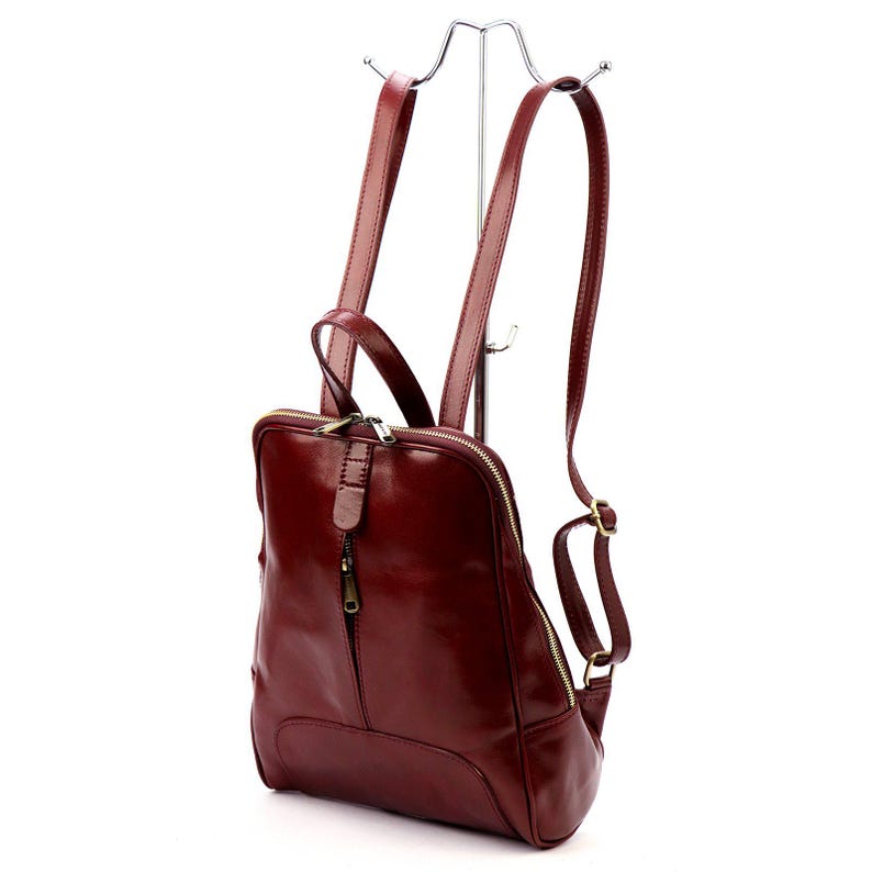 May include: A burgundy leather backpack with adjustable straps and a top handle. The bag features a zippered main compartment and a smaller front pocket. The hardware is gold-toned, and the bag is displayed on a silver metal stand.