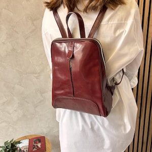 May include: A burgundy leather backpack with a square shape and a top handle. The backpack has a vertical zipper and a smaller zippered pocket on the side. The straps are adjustable. The bag is worn over a white shirt.
