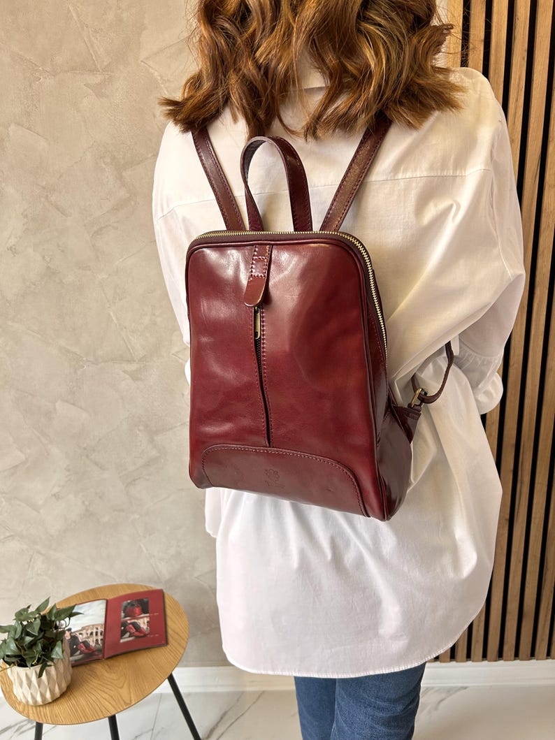 May include: A burgundy leather backpack with a square shape and a top handle. The backpack has a vertical zipper and a smaller zippered pocket on the side. The straps are adjustable. The bag is worn over a white shirt.