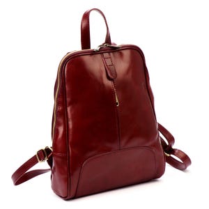 May include: A burgundy leather backpack with a glossy finish. The bag features a top handle, adjustable straps, and a gold-toned zipper. The design includes a front pocket and a small leather flap with a zipper pull.