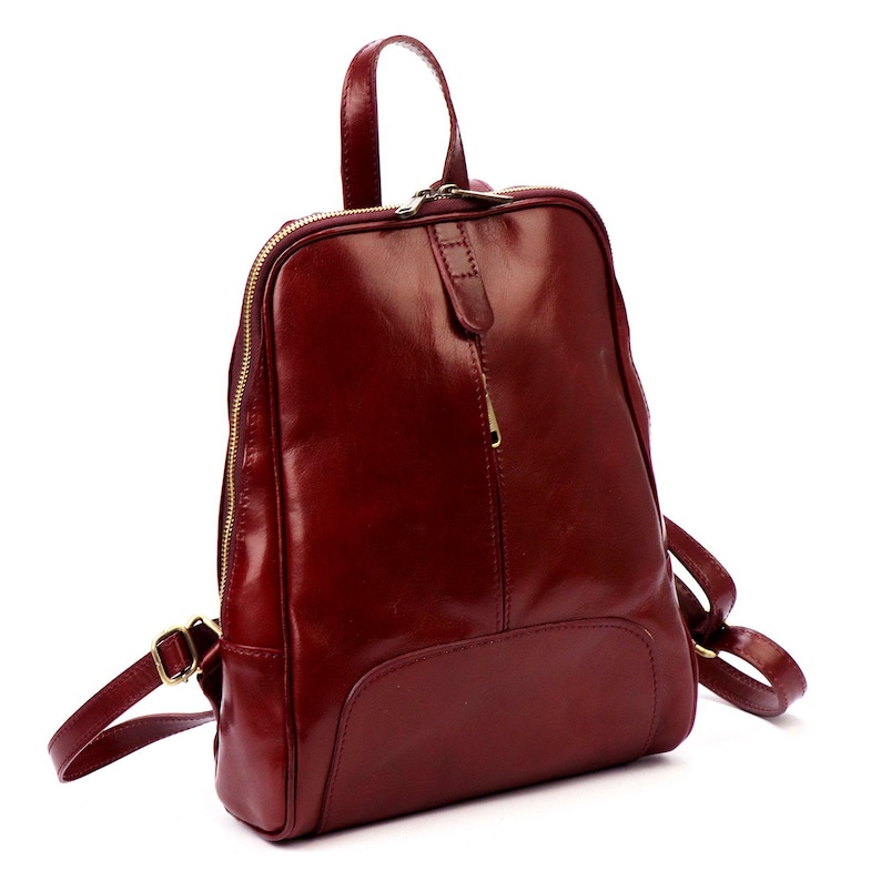 May include: A burgundy leather backpack with a glossy finish. The bag features a top handle, adjustable straps, and a gold-toned zipper. The design includes a front pocket and a small leather flap with a zipper pull.