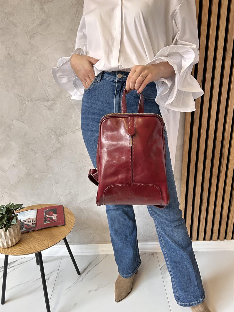 May include: A burgundy leather backpack with a top handle and zipper closure. The bag has a rectangular shape with rounded corners. The person is wearing blue jeans and a white blouse with ruffled sleeves. A small side table with a book and plant is visible.
