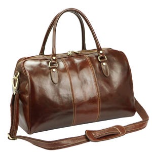 Puede incluir: Bolso de viaje de cuero marr&oacute;n brillante con correa de hombro desmontable y dos asas superiores. El bolso presenta detalles dorados, incluyendo la cremallera y las fijaciones de la correa. Dise&ntilde;o cl&aacute;sico y adecuado para viajes, con un aspecto pulido.