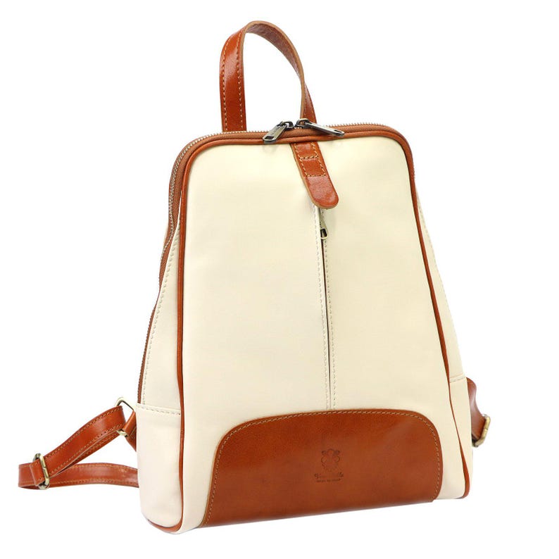 May include: Cream and brown leather backpack with a top handle and adjustable straps. The backpack features a cream-colored main body with brown trim and a brown bottom panel. A zipper runs along the top and side for access to the interior.
