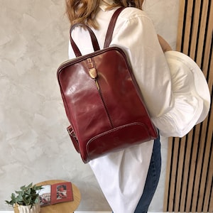May include: A burgundy leather backpack with a rectangular shape and a top handle. The backpack has a front zipper and adjustable shoulder straps. The bag is worn over the shoulder.