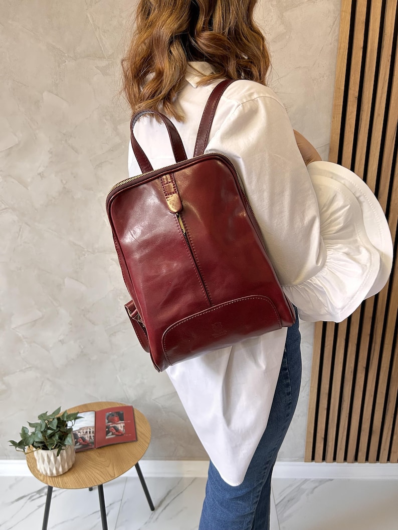 May include: A burgundy leather backpack with a rectangular shape and a top handle. The backpack has a front zipper and adjustable shoulder straps. The bag is worn over the shoulder.