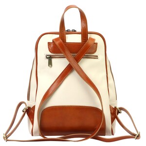 May include: Cream and brown leather backpack with a top handle and adjustable shoulder straps. The backpack features a front pocket and a zippered main compartment. The straps cross in an X shape on the back.