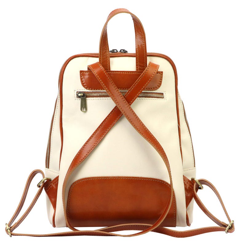 May include: Cream and brown leather backpack with a top handle and adjustable shoulder straps. The backpack features a front pocket and a zippered main compartment. The straps cross in an X shape on the back.