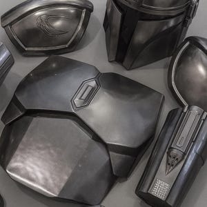 May include: Metallic gray Mandalorian armor pieces, including a helmet, chest plate, shoulder pads, and gauntlets. The armor has a weathered appearance with detailed paneling and a sleek, futuristic design. The pieces are arranged on a gray surface.