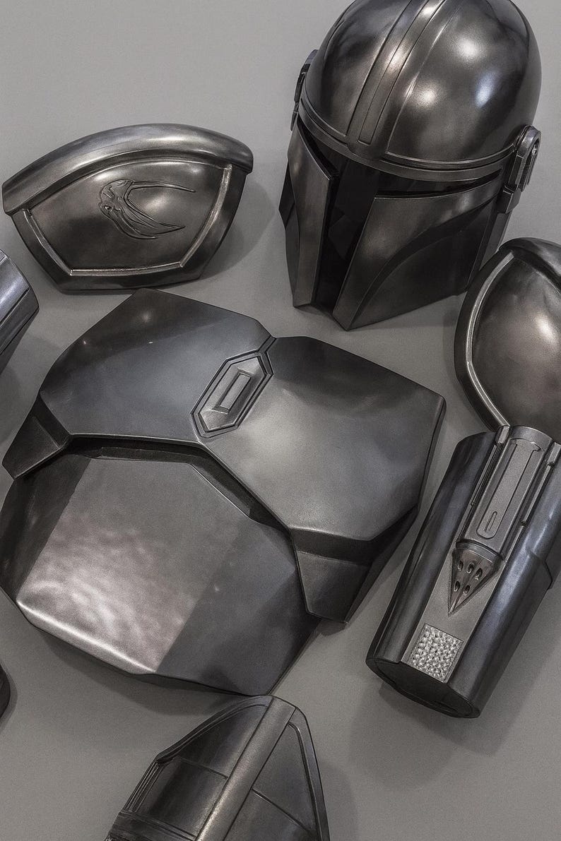 May include: Metallic gray Mandalorian armor pieces, including a helmet, chest plate, shoulder pads, and gauntlets. The armor has a weathered appearance with detailed paneling and a sleek, futuristic design. The pieces are arranged on a gray surface.