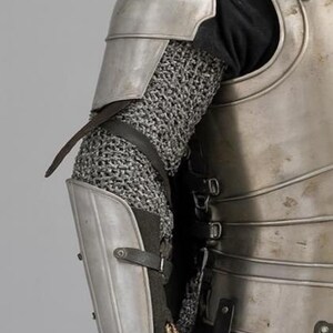 May include: Detailed view of medieval armour, featuring a silver metal shoulder plate, chainmail sleeve, and lower arm guard. The armour is fastened with leather straps and buckles, indicative of a historical or fantasy setting.