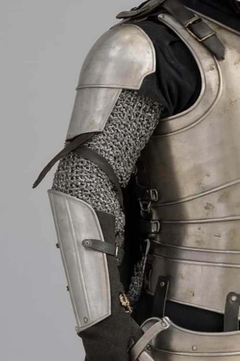 May include: Detailed view of medieval armour, featuring a silver metal shoulder plate, chainmail sleeve, and lower arm guard. The armour is fastened with leather straps and buckles, indicative of a historical or fantasy setting.