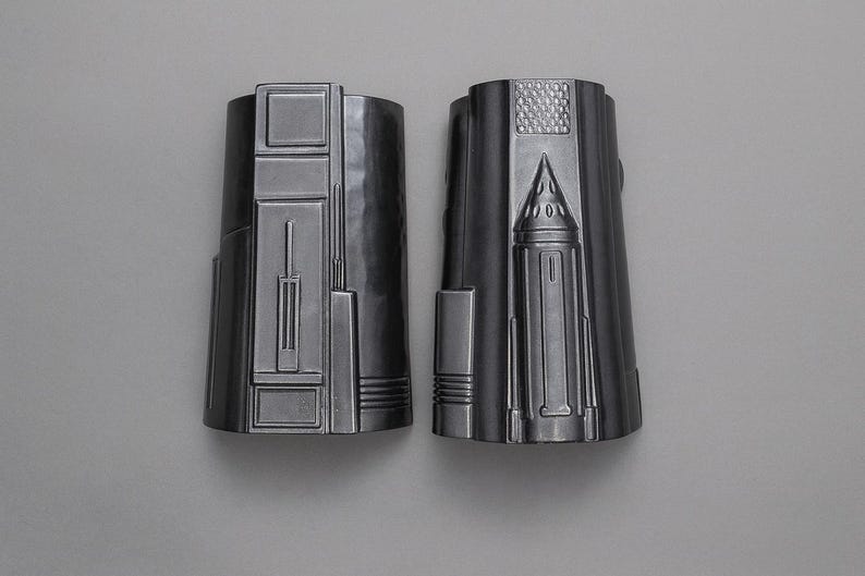 May include: Two silver-colored, cylindrical objects with detailed, geometric designs. The objects have a metallic sheen and are set against a neutral background. The design includes rectangular and triangular shapes.