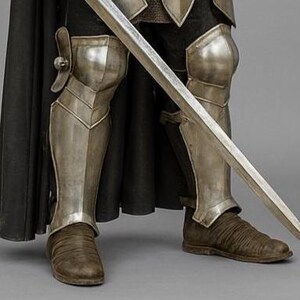 May include: Close-up of a knight's lower body, clad in silver armour and brown boots. A long, silver sword is held diagonally across the legs. A black cape drapes behind the armour, creating a dramatic contrast.