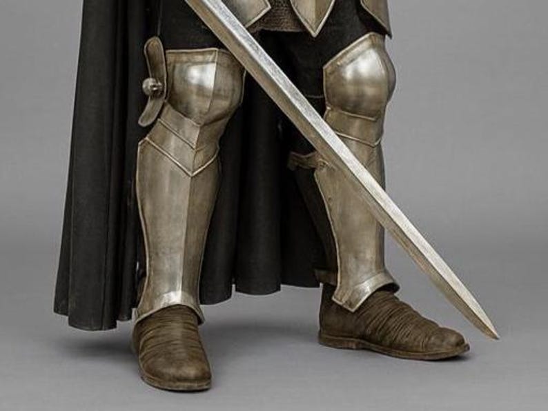 May include: Close-up of a knight's lower body, clad in silver armour and brown boots. A long, silver sword is held diagonally across the legs. A black cape drapes behind the armour, creating a dramatic contrast.