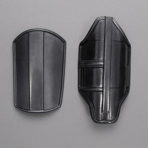 May include: Two dark gray metal arm guards. One is a rounded rectangle with vertical lines, the other is a contoured shape with horizontal bands and vertical lines. The guards are displayed against a gray background.
