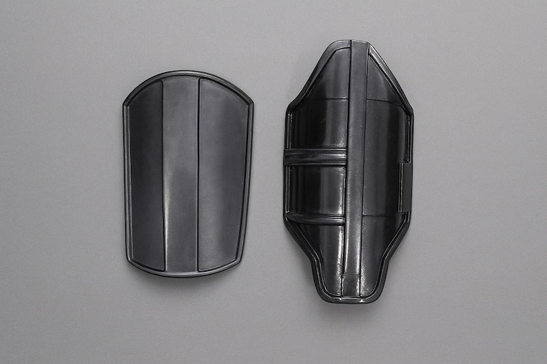 May include: Two dark gray metal arm guards. One is a rounded rectangle with vertical lines, the other is a contoured shape with horizontal bands and vertical lines. The guards are displayed against a gray background.