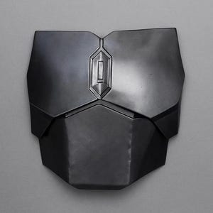 May include: A dark gray chest plate with a geometric design. The plate has a central, raised rectangular detail and is composed of multiple angular sections. The surface has a metallic sheen, suggesting a protective garment or armor.