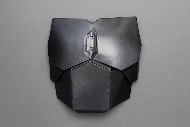 May include: A dark gray chest plate with a geometric design. The plate has a central, raised rectangular detail and is composed of multiple angular sections. The surface has a metallic sheen, suggesting a protective garment or armor.