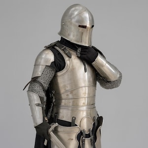 May include: A complete set of medieval armour, including a helmet, chest plate, arm guards, and chainmail. The armour is made of polished metal with intricate details. The person is wearing black gloves and a black shirt.