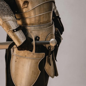 May include: A detailed view of medieval armour, featuring a silver breastplate, shoulder protection, and leg guards. The armour is riveted and worn with black gloves. A sword with a silver blade and black handle is held in the gloved hand.