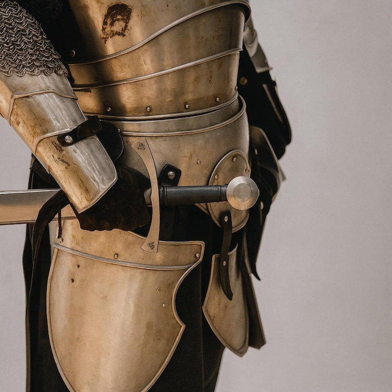 May include: A detailed view of medieval armour, featuring a silver breastplate, shoulder protection, and leg guards. The armour is riveted and worn with black gloves. A sword with a silver blade and black handle is held in the gloved hand.