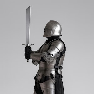 May include: A person in full medieval armour, including a helmet, shoulder guards, and chest plate, holding a large, silver sword. The armour is detailed with chainmail and straps. The background is a solid, neutral grey.