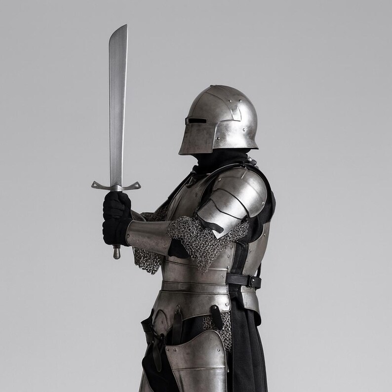 May include: A person in full medieval armour, including a helmet, shoulder guards, and chest plate, holding a large, silver sword. The armour is detailed with chainmail and straps. The background is a solid, neutral grey.