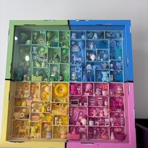 Puede incluir: Una caja expositora cuadrada dividida en cuatro cuadrantes de colores verde, azul, amarillo y rosa. Cada sección contiene una colección de figuras en miniatura, juguetes y objetos pequeños coordinados por color con el fondo de su compartimento.