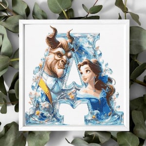 May include: A framed cross-stitch depicting the letter "A" with images of Belle and the Beast from Beauty and the Beast. The design uses blue, yellow, and white, with floral details. The frame is white, set against a white surface with green foliage.