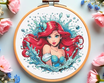 May include: A cross-stitch embroidery of a cartoon character with red hair and a blue top, surrounded by water splashes. The embroidery is set in a wooden hoop, with pink and blue flowers around the edge.