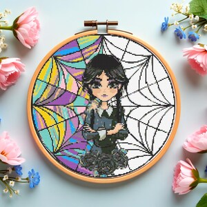 May include: A cross-stitch embroidery hoop featuring a cartoon character with black hair and a dark dress. The background is split, with a colorful stained-glass design on one side and a spiderweb pattern on the other. The wooden hoop is surrounded by pink and blue flowers.